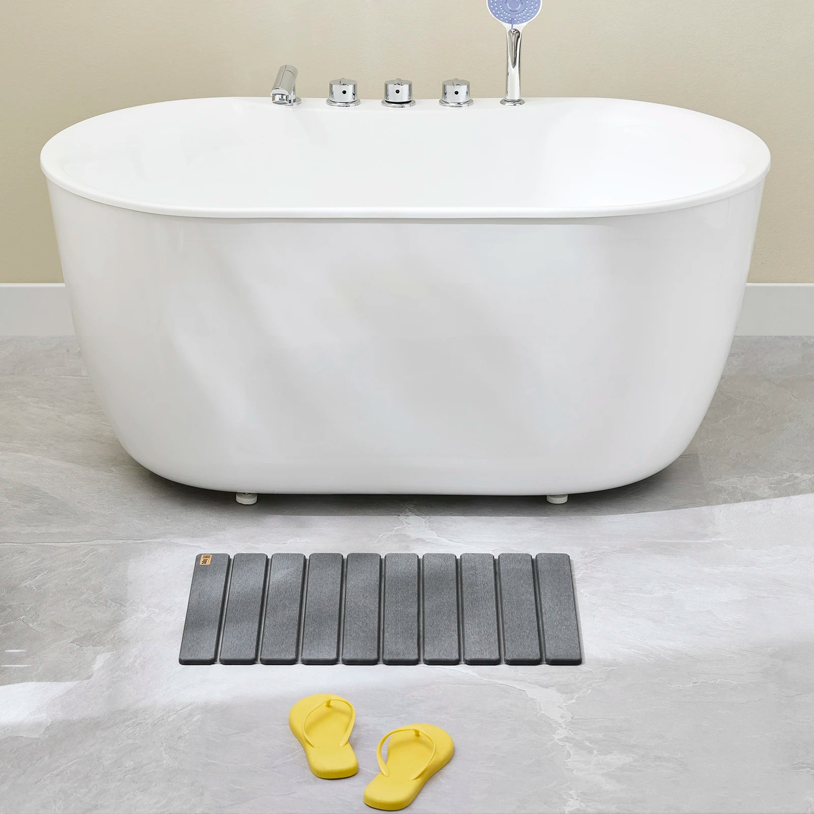 Tapis de bain rectangulaire gris positionné devant une baignoire sur un carrelage clair.