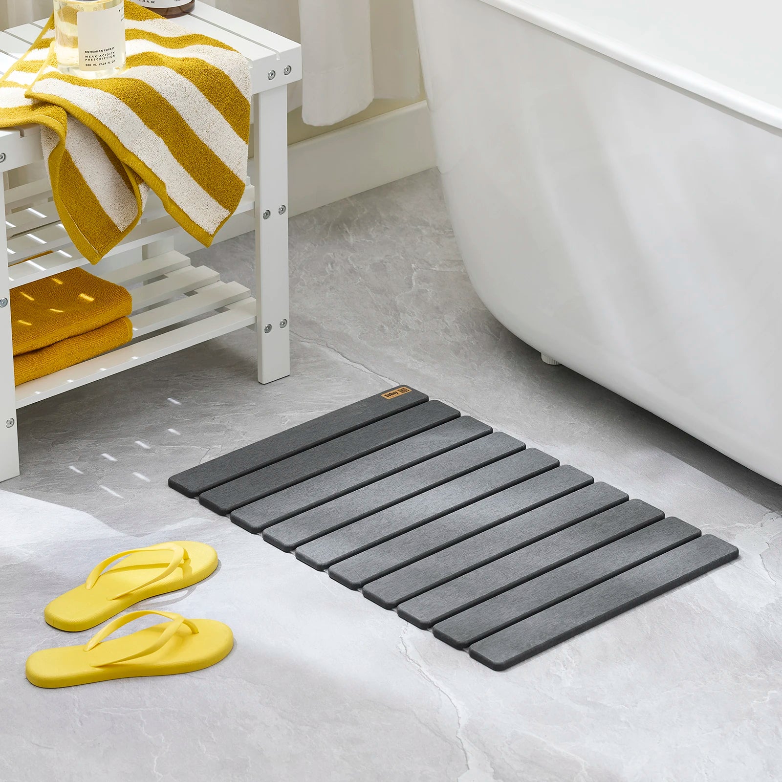 Tapis de bain gris placé sur le sol d’une salle de bain près d’une baignoire avec des sandales jaunes.